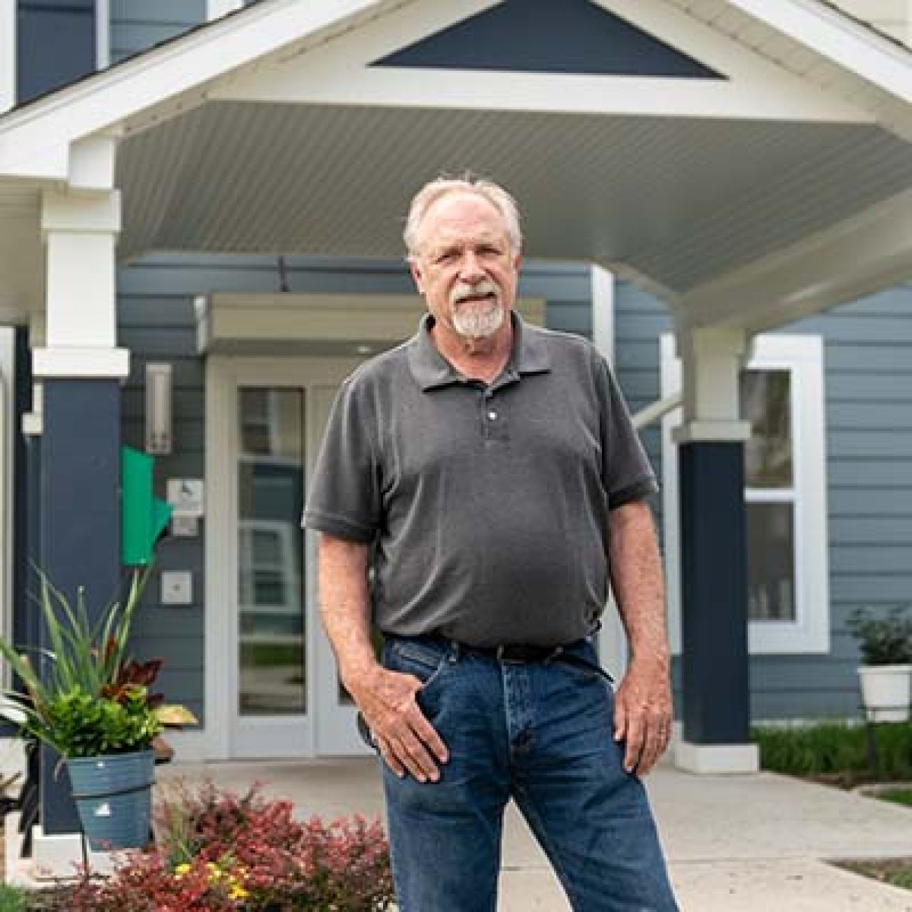 Mike Hirst is wearing a grey polo shirt and jeans. He is posing for a picture in front of a building