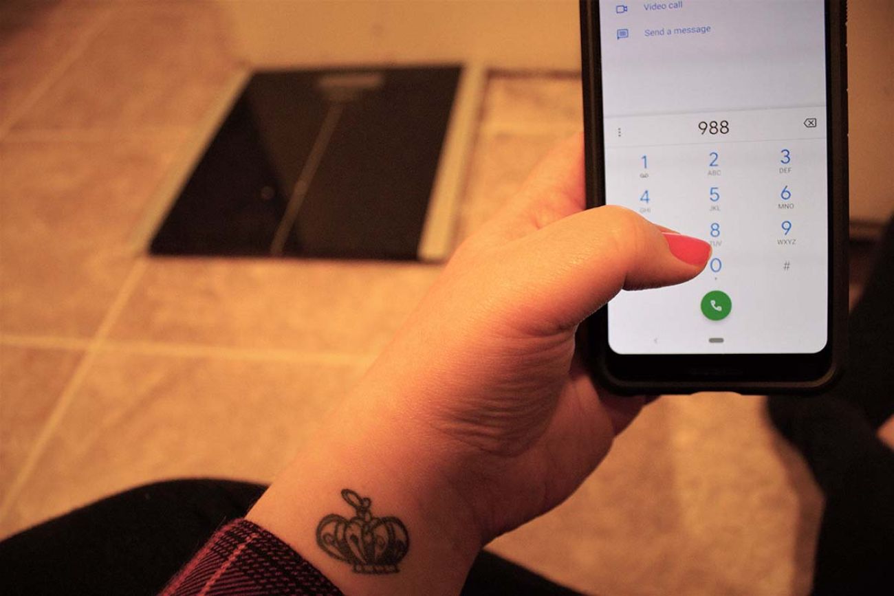 A woman with a tattoo dials the national suicide hotline from its new 988 number from the floor in front of her scale. 