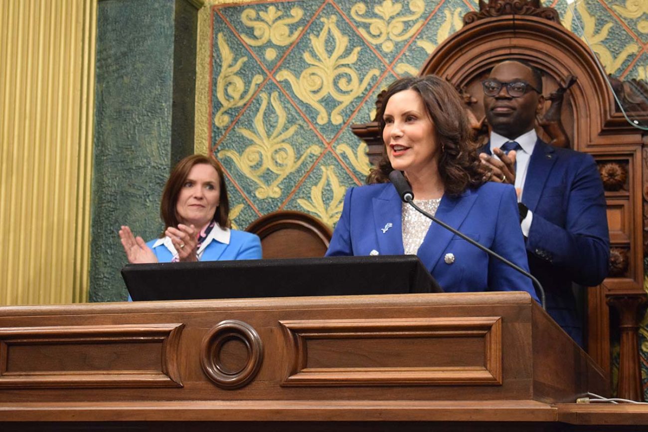 Gov. Gretchen Whitmer speaking into a microphone. Winnie Brinks and Garlin Gilchrist are in the background