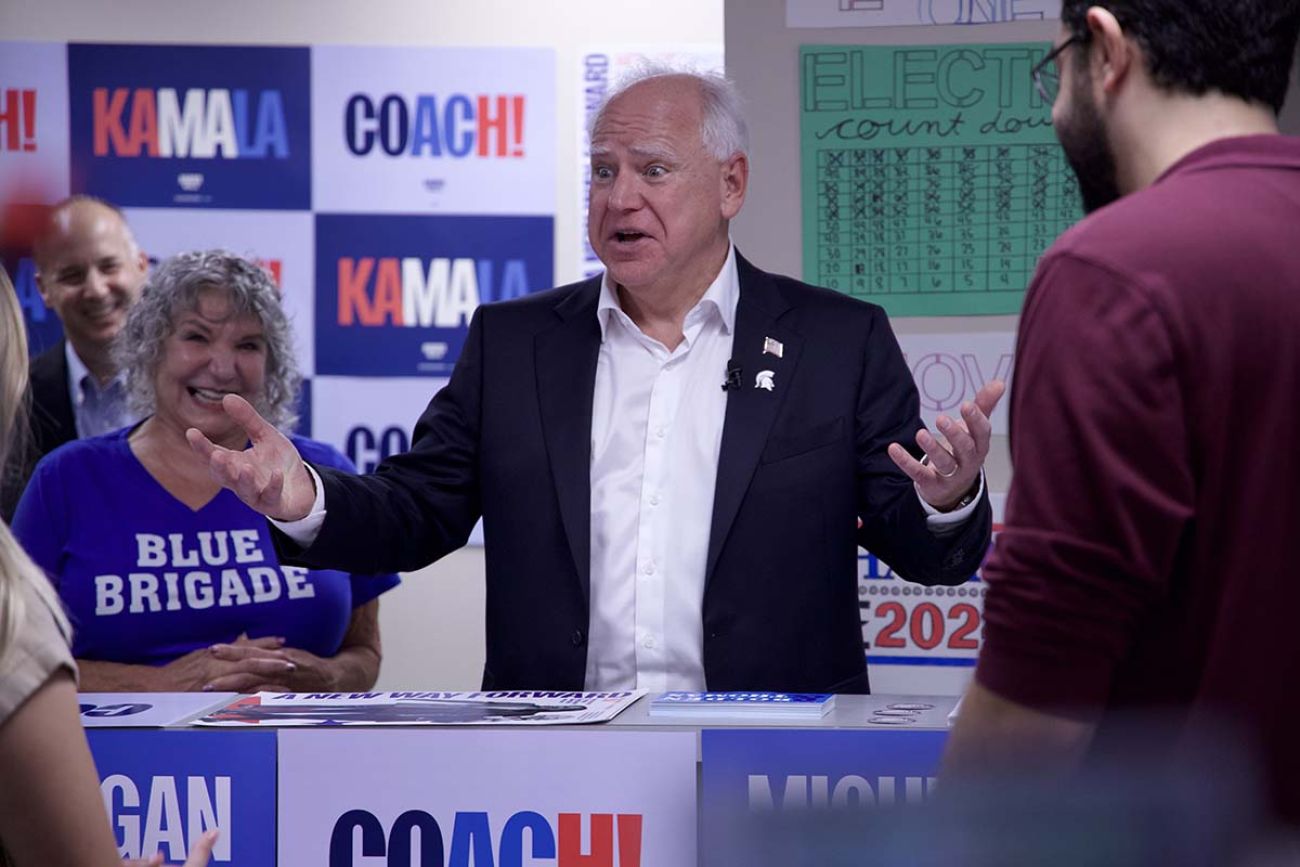 Tim Walz is wearing a white shirt and navy jacket. There are pro-Harris signs behind him