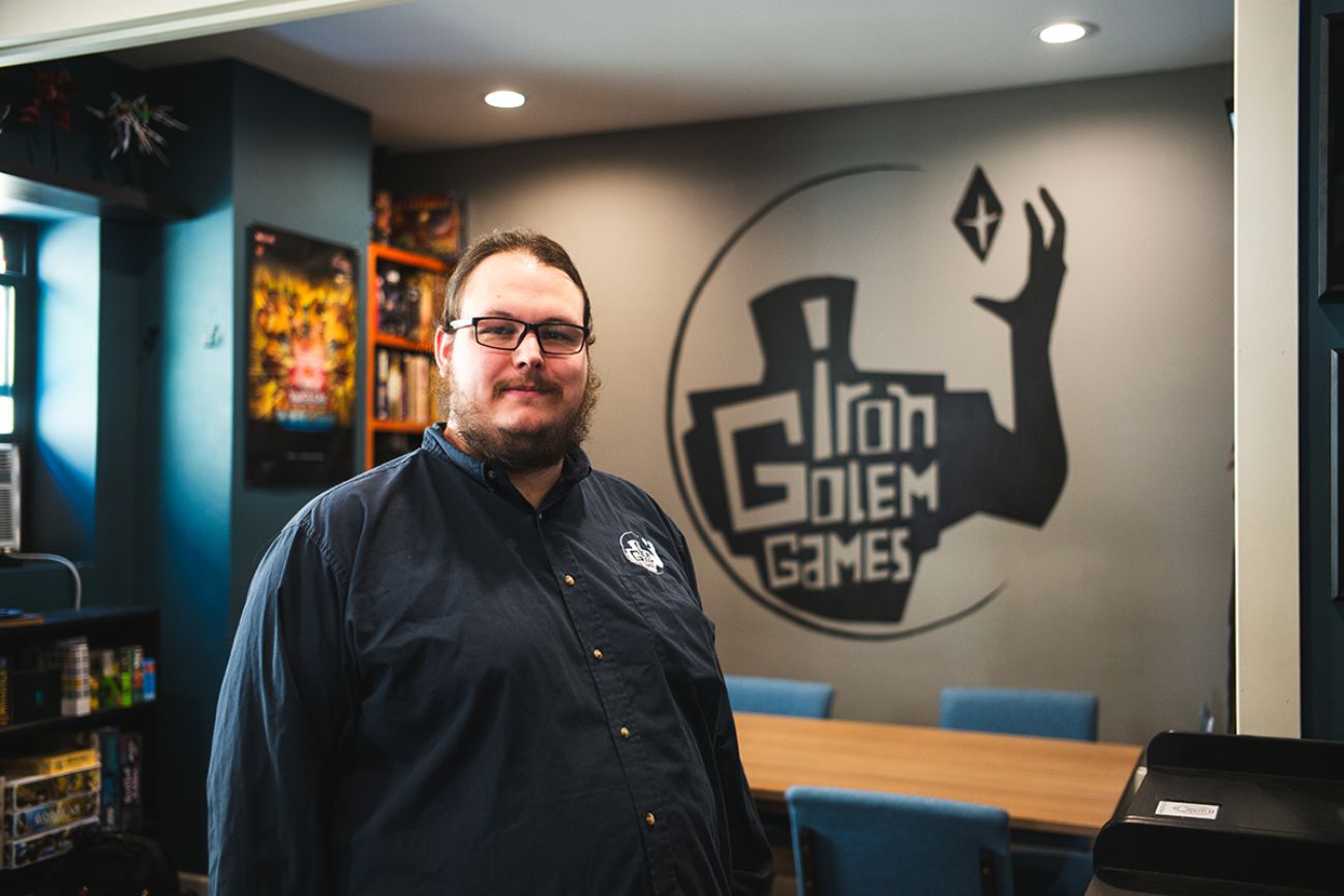 Joseph Baker wears a long-sleeve dark-colored shirt with the Iron Golem Games logo. He is standing in Iron Golem Games in Marquette