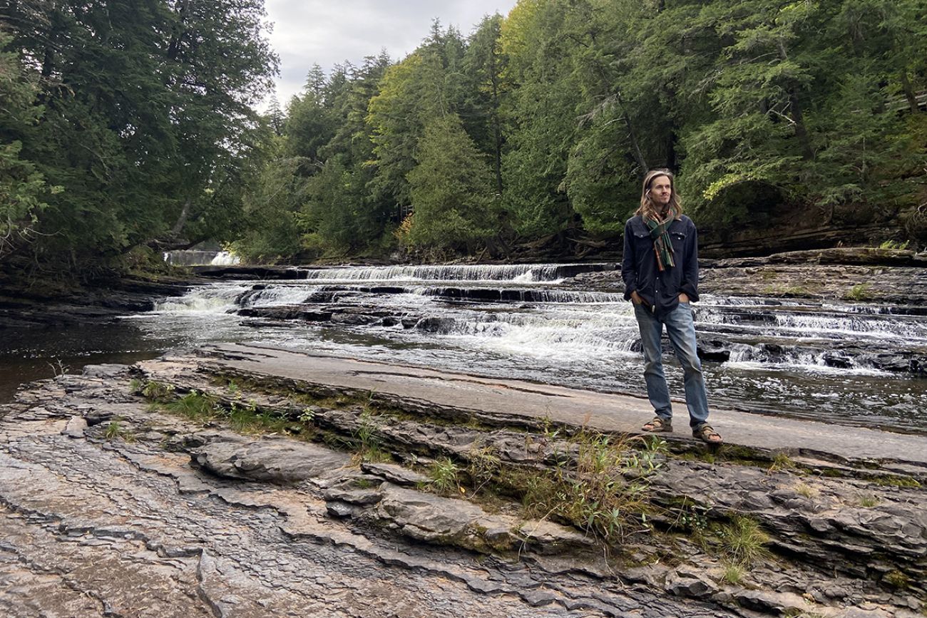 Tom Grotewohl standing next a river. He is wearing a scraf, blue long sleeve shirt and jeans