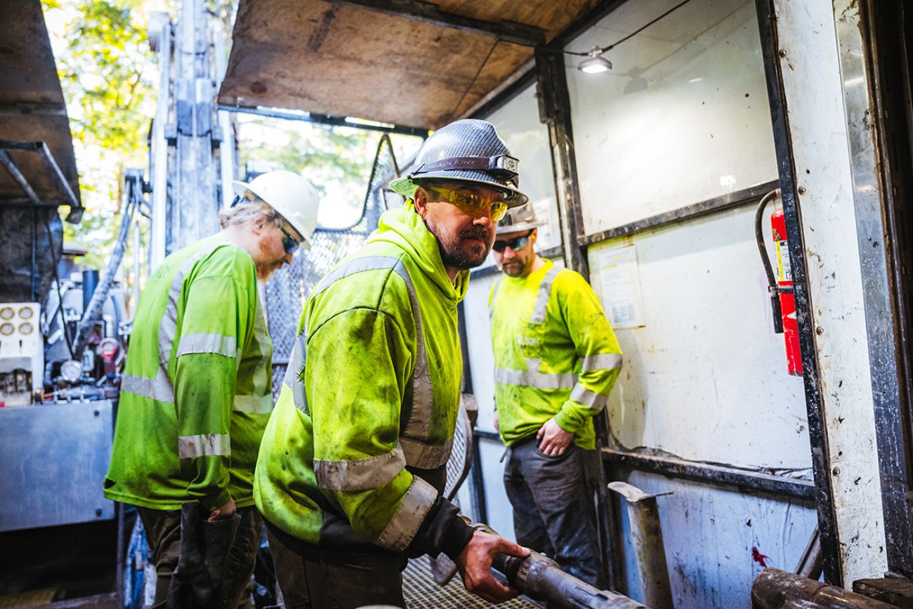 Three men wearing green vests perate a core drilling rig deep 