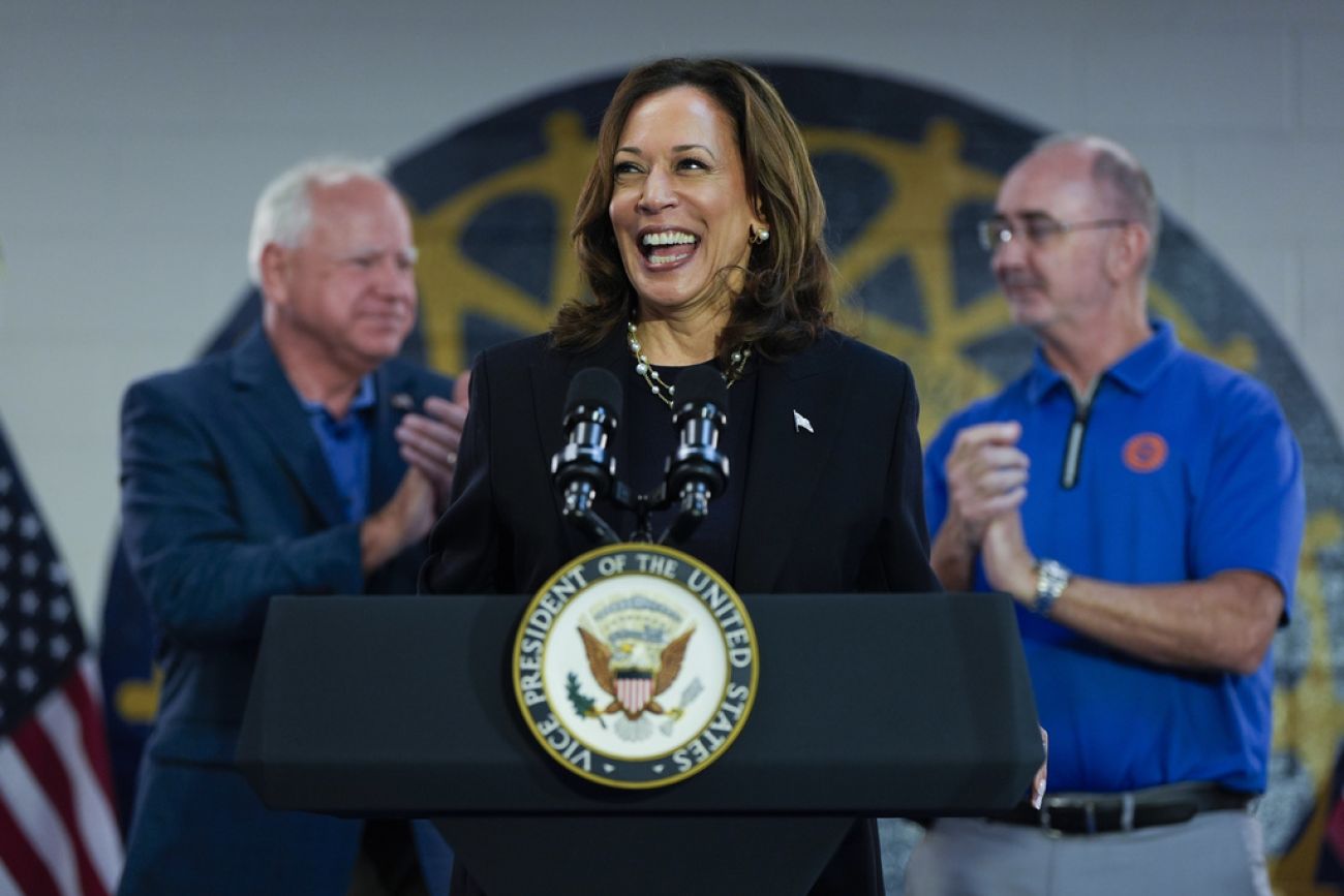 Kamala Harris stands in front of a podium. Minnesota Gov. Tim Walz, left, and UAW President Shawn Fain are in the background