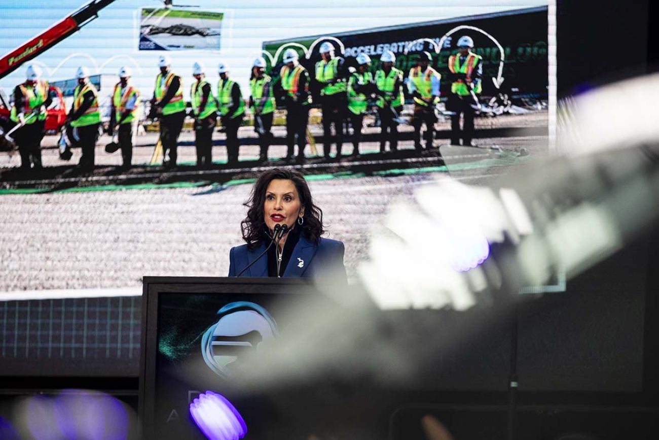 Gov. Gretchen Whitmer on stage. Behind her is a picture of people breaking ground on a project.