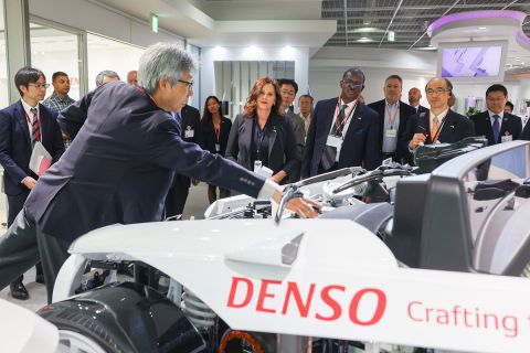 Gov. Gretchen Whitmer surrounded by many other people looking at a white Denso vehicle