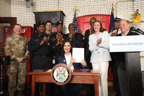 Gov. Gretchen Whitmer holds up the signed budget. She is in a firehouse with people surrounding her, clapping