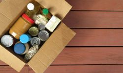 A box of donated canned goods and non-perishable foods for a food pantry for the poor sitting on red, wood panel background. 