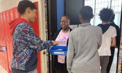 a teacher holding a blue bin and kids putting their cellphones in there